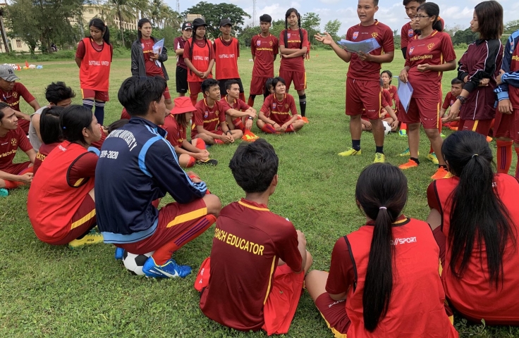 A group of young soccer players are gathered in a circle. They are in red and yellow uniforms. Half are sitting down with their backs to the camera.