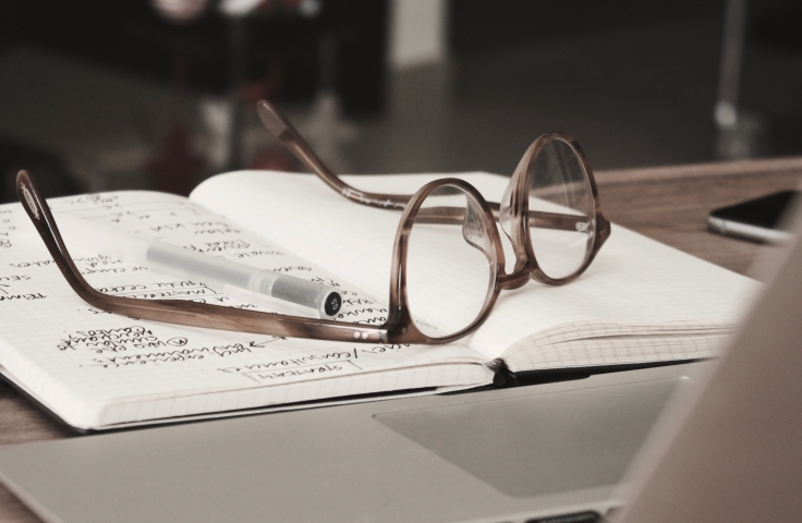 An open notebook sits on a table. There is writing on the left page. A pair of spectacles sits on top of the notebook.