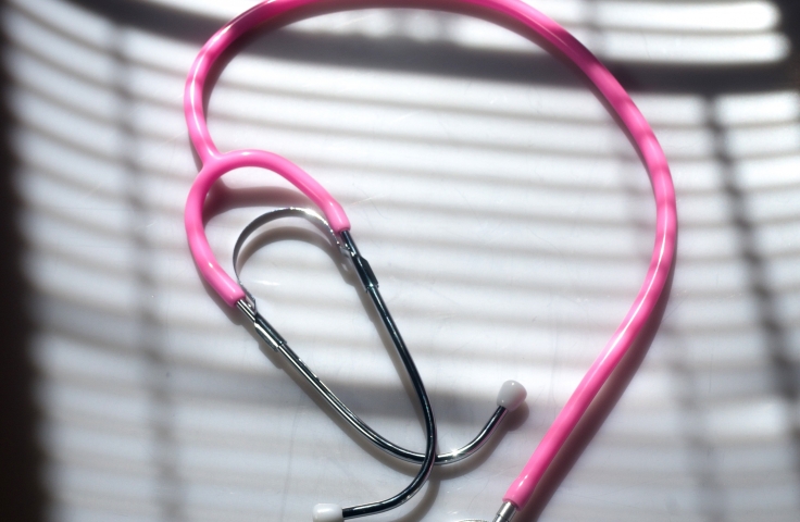 A pink stethoscope is sitting on a table. An open window with blinds is casting a shadow over the image.